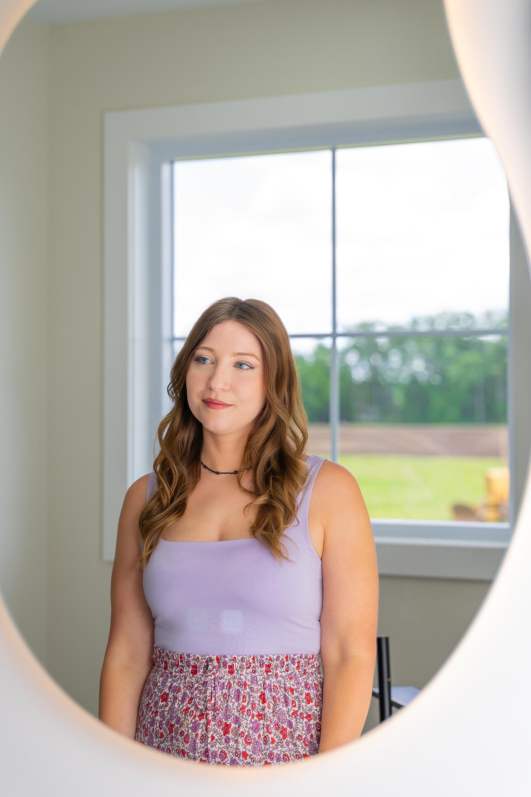 Reflection of a woman in an oval, illuminated mirror. She has wavy brown hair, wears a purple tank top & floral skirt, and looks left.