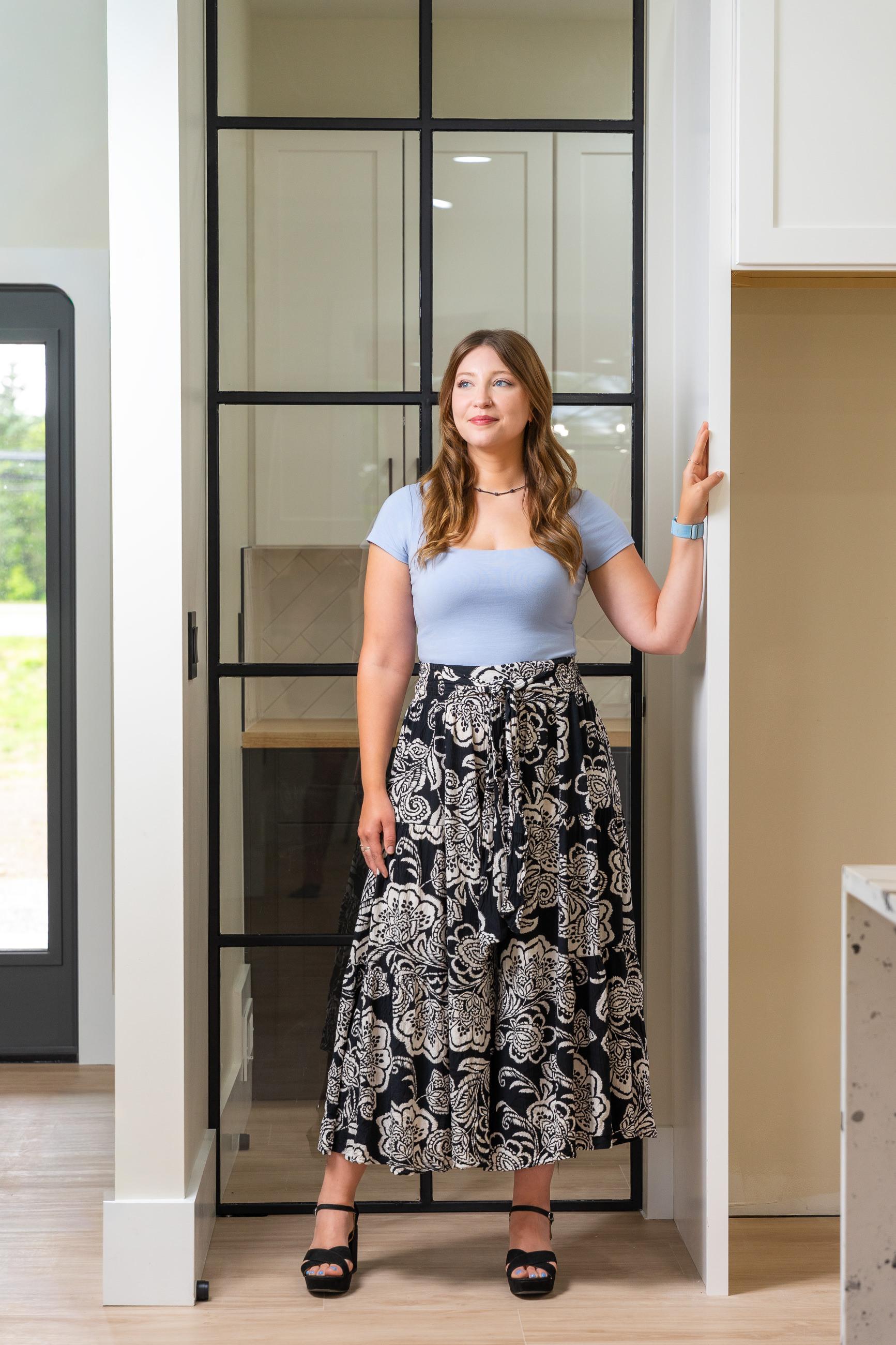 A woman in a light blue top, black and white floral maxi skirt, and heels stands in a modern interior with a black grid glass partition.