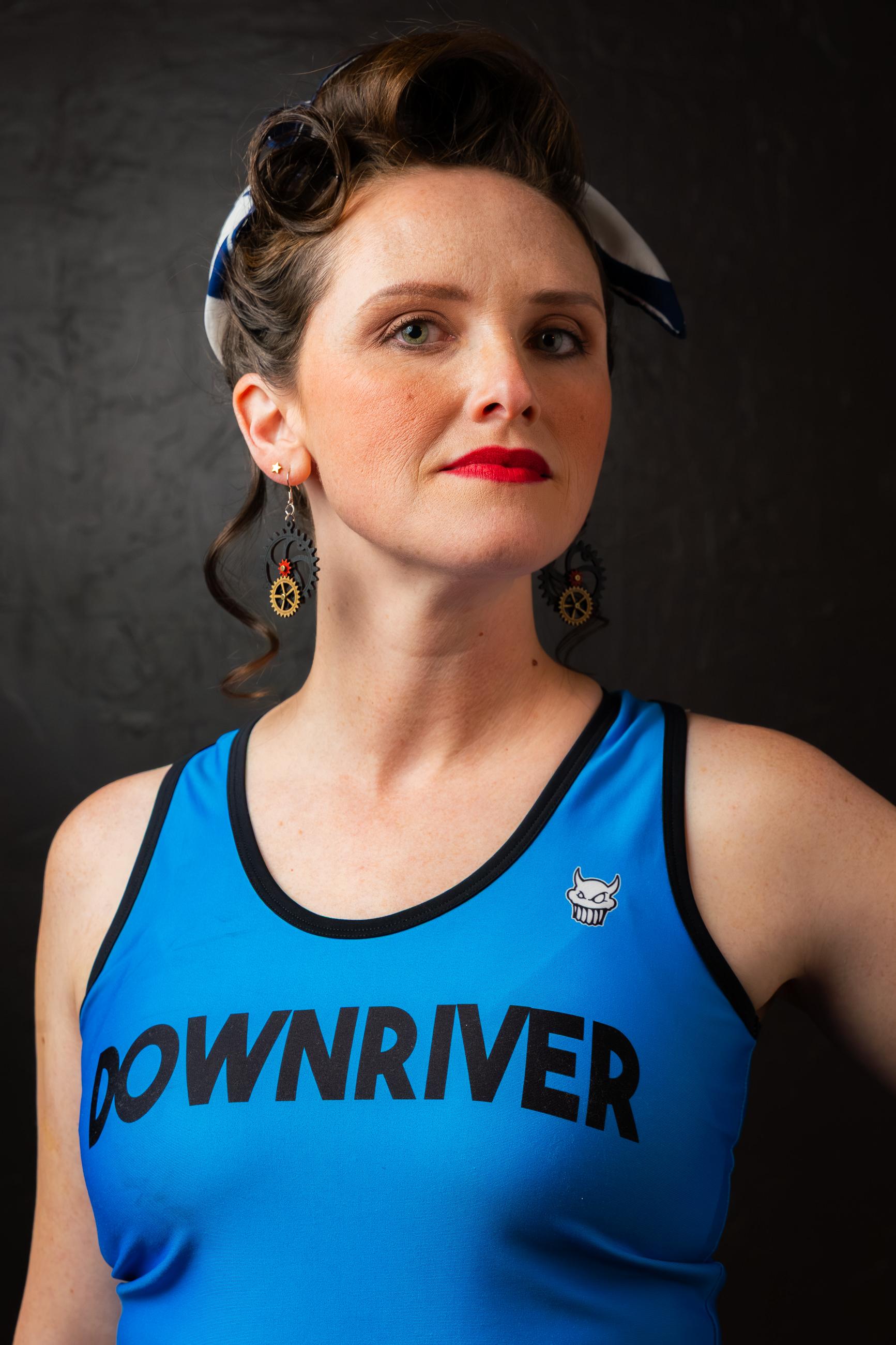 A woman with a retro hairstyle and bandana wears a blue "DOWNRIVER" tank top, red lipstick, and steampunk gear earrings against a dark background.