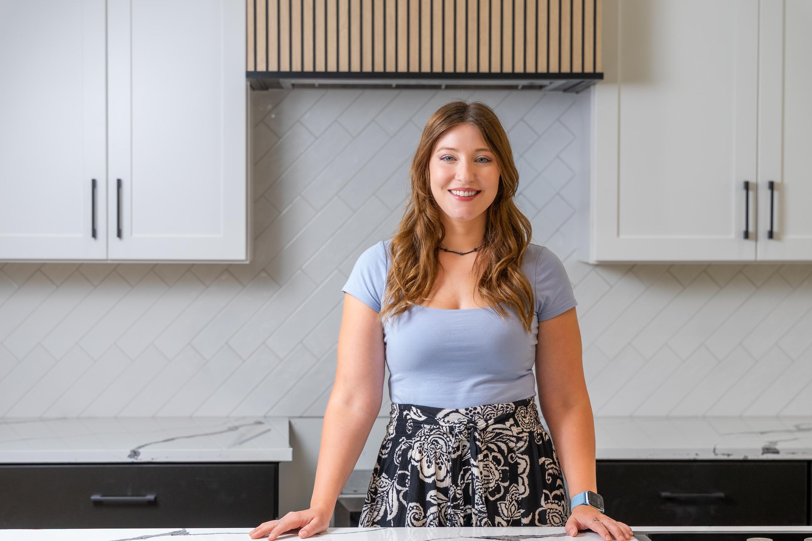 Photograph of a woman in a blue top and floral skirt, smiling in a kitchen