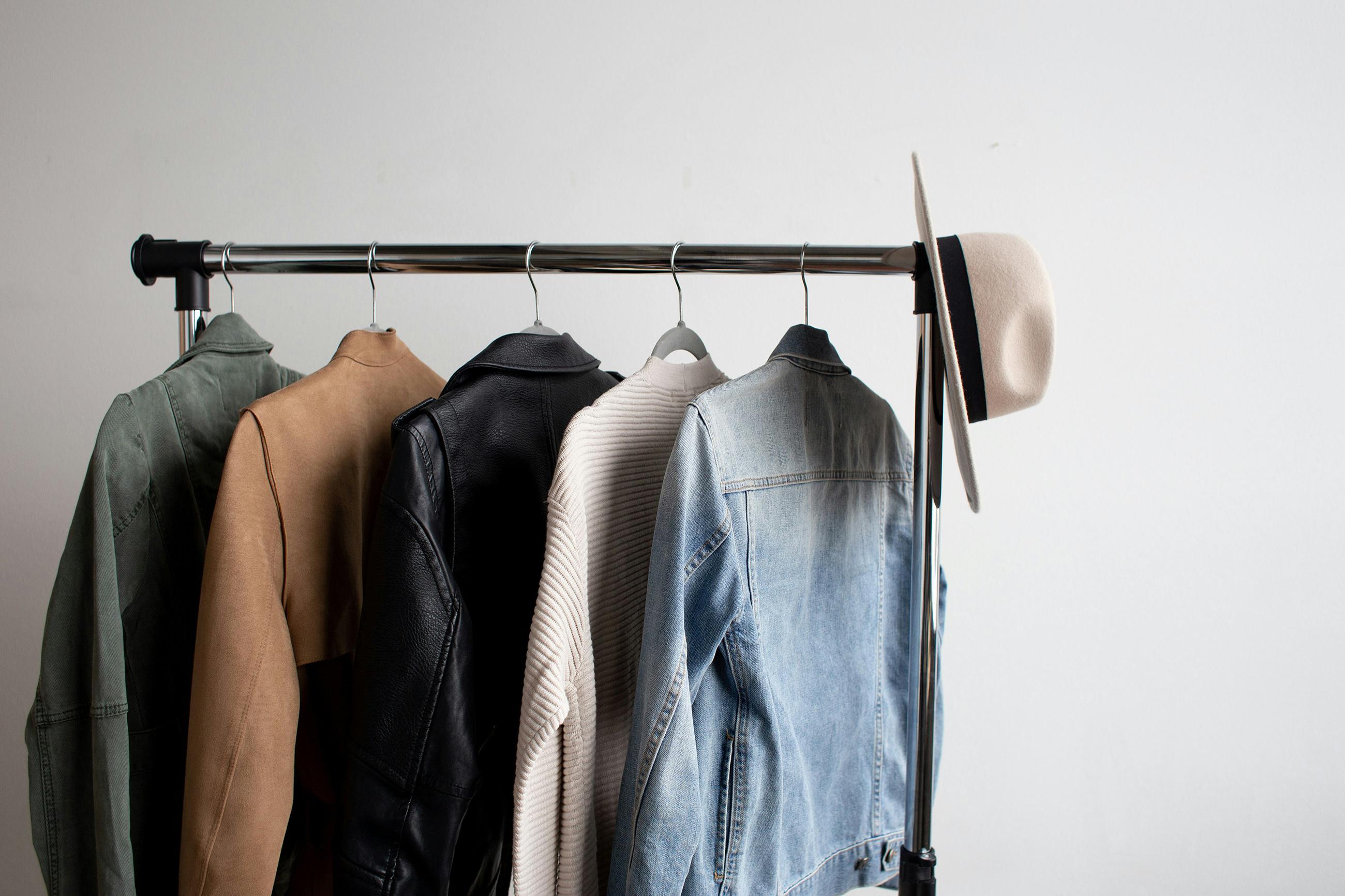 A metal clothing rack holds a green jacket, tan jacket, black leather jacket, light sweater, denim jacket, and a beige fedora hat.