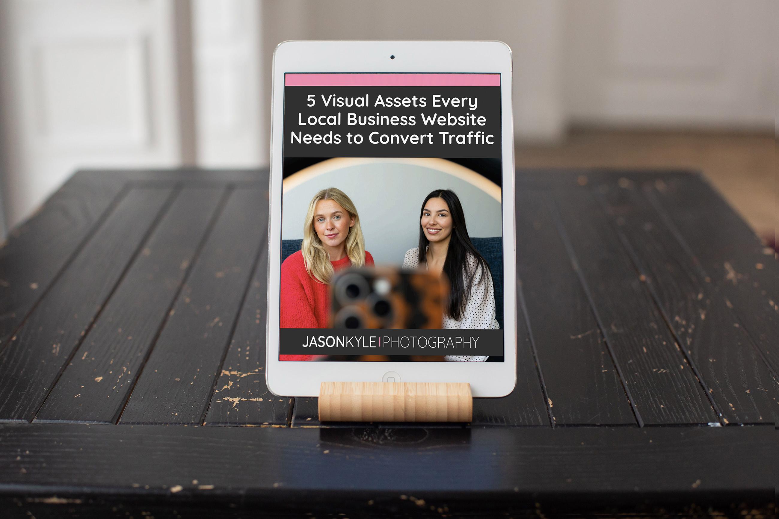 An iPad displays an article titled "5 Visual Assets Every Local Business Website Needs to Convert Traffic" with photos of two women.
