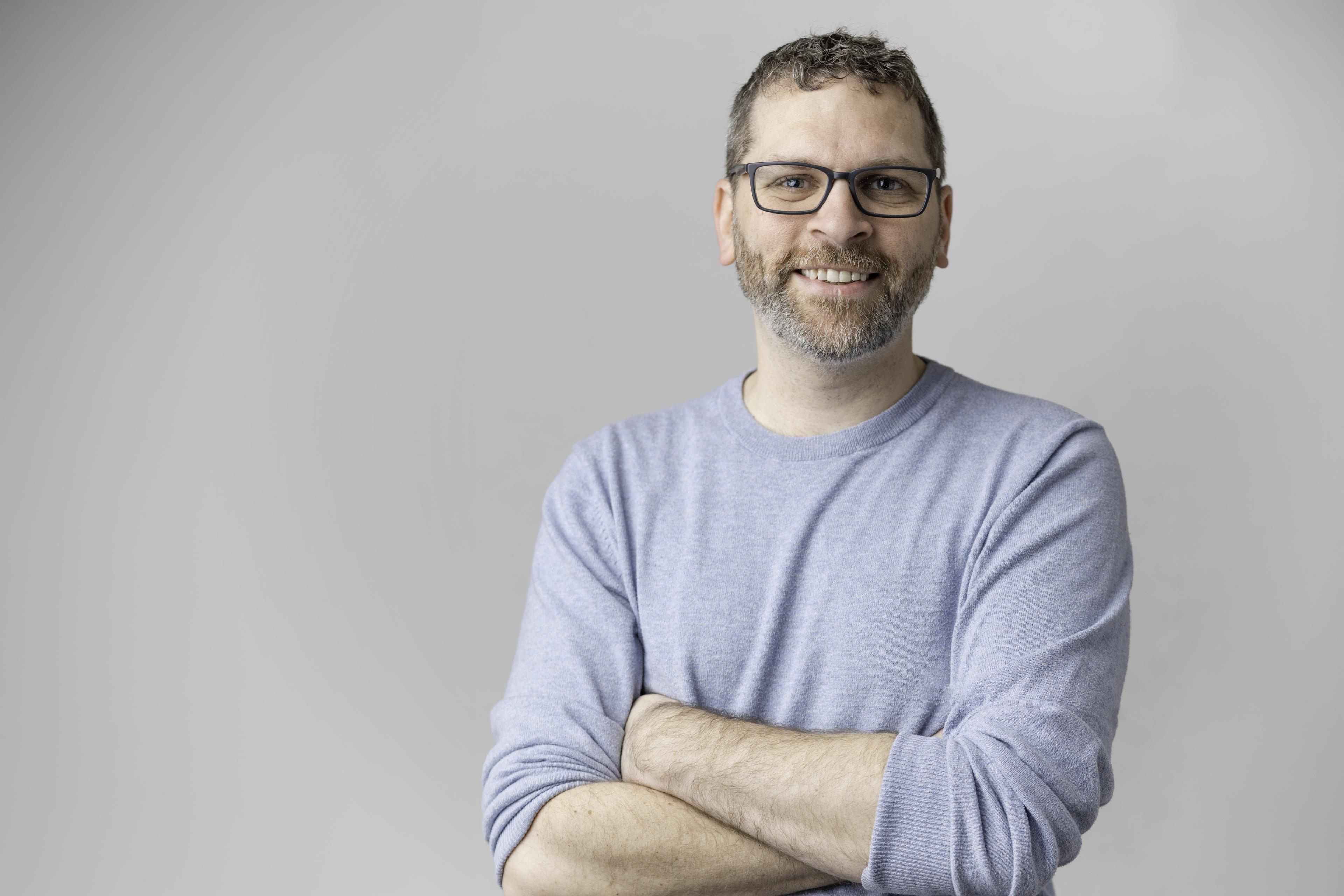 A smiling man with a beard and dark-rimmed glasses wears a light blue sweater and crosses his arms against a plain light gray background.